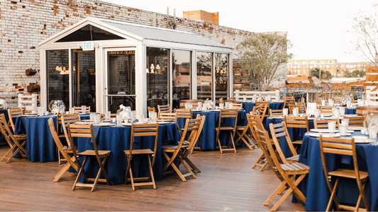Rooftop event space with round tables set in navy blue linens and wooden folding chairs, string lights glowing inside a glass-walled pavilion, exposed brick and downtown Oklahoma City skyline in the background.
