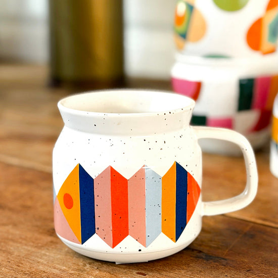 A white ceramic mug with a colorful geometric pattern of orange, pink, blue, and yellow shapes, sitting on a rustic wooden table with other mugs blurred in the background.