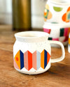 A white ceramic mug with a colorful geometric pattern of orange, pink, blue, and yellow shapes, sitting on a rustic wooden table with other mugs blurred in the background.