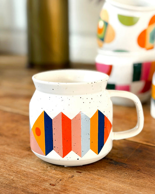 A white ceramic mug with a colorful geometric pattern of orange, pink, blue, and yellow shapes, sitting on a rustic wooden table with other mugs blurred in the background.