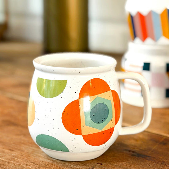 A white ceramic mug with a colorful mid-century modern geometric pattern, sitting on a rustic wooden table with other mugs blurred in the background.