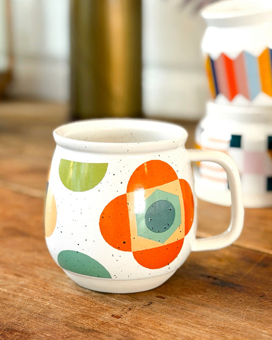 A white ceramic mug with a colorful mid-century modern geometric pattern, sitting on a rustic wooden table with other mugs blurred in the background.