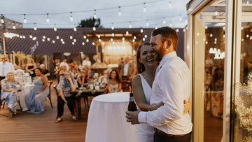 Couple embracing at an outdoor event with string lights and people in the background